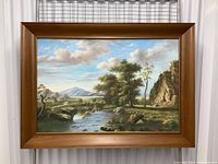 Framed oil painting showing full landscape scene with river, trees, rocks, mountains, and sky.