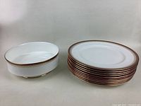 Photo showing a stack of fine bone china dinner plates and one serving bowl placed beside them. Plates have detailed gold and reddish-brown border with gold rim.