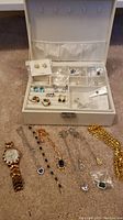 White jewelry box open showing multiple pairs of earrings inside and various items on carpet