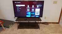 TCL Roku TV powered on, screen showing Roku home interface, placed on brown metal/plastic stand in carpeted room