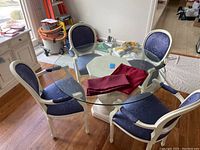 Full view of round glass dining table with four blue upholstered chairs around it. Two burgundy tablecloths lying on the table.
