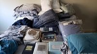 Photo showing various folded bedding sets, pillows, and blankets stacked on the floor in different colors including blue, gray, and neutral tones.