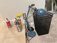 Photo showing blue adjustable office lamp, black metal mesh trash can, cup with assorted writing instruments, and office duster spray bottle on tiled floor.