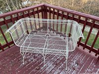 White metal wire loveseat on a red wooden deck with visible weathering and surface dirt.