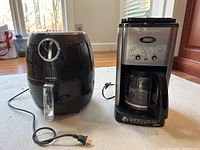 Black Gourmia air fryer and stainless steel Cuisinart coffee maker placed side by side on a wooden floor.
