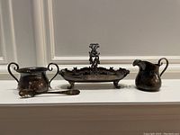 Front view of all three sterling silver serving pieces together: an elongated handled dish with ornate divider, a creamer jug, and a sugar bowl with spoon.