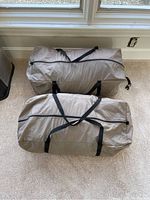 Two gray large duffel bags with black straps placed indoors on carpeted floor near window, front and top view visible.