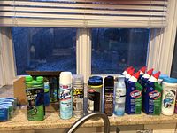 Photo showing array of cleaning products on kitchen counter by window including Lysol spray, Flex Seal clear, Woodman wood polish, Clorox toilet cleaner, Glade carpet cleaner, 3M office cleaner.