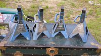 Photo showing four blue and black metal axle stands placed outdoors on a wooden surface. Each stand has a triangular base and adjustable height mechanism with handles.