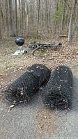 Two large black chain link fence rolls placed on ground outdoors, in a natural wooded setting with dry leaves and gravel. Nearby is a small black kettle grill and some metal frames partially visible in the background.