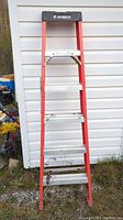 Side view of 6 ft Featherlite fiberglass step ladder against white siding, showing red frame, metal steps, black top cap, and Featherlite branding.