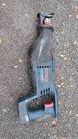 Bosch reciprocating saw body only in used condition, showing the side with the Bosch logo and red trigger, placed on a ground surface with leaves and dirt.
