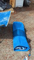 Folded blue camping cot inside a blue carry bag with black straps, placed outdoors on a gravel surface.