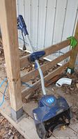 Photo of a blue and grey Snowjoe electric snow sweeper resting against a wooden fence outdoors with leaves on the ground.