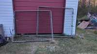 Four metal chain-link fence gates leaning against a red wall and white building exterior on grass ground. Gates have tubular metal frames and chain-link mesh, with visible hinges and latch hardware.