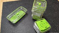 Stacked clear rectangular plastic vacuum sealing containers fitted with green lids and air valves, each containing green perforated inserts on a black countertop.