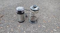 An electric coffee grinder and a glass French press placed outdoors on asphalt, both used and showing signs of wear.