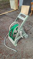 Photo of gray Ames Reel Easy hose reel with green garden hose and white hose connected, placed on gravel surface outdoors with some other outdoor items in background.