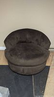 Large brown swivel chair with rounded back and thick cushion, placed against a beige wall on mixed flooring.