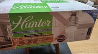 Boxed 52-inch Hunter ceiling fan showing image of fan, brand Hunter, mentions Surespeed motor technology, and product details in multiple languages.