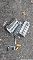 Chrome-plated pasta roller and cutter attachments with crank handle and clamp lying on asphalt surface.