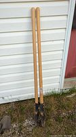 Photo of two post hole diggers with wooden handles leaning against a white siding wall