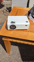 White compact projector placed on wooden table outdoors, showing front lens and controls on top.