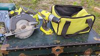 Photo showing a Ryobi circular saw and drill next to a yellow and black carrying bag on a worn green trunk.