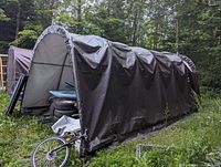 Side view of brown tarp covered temporary shelter with closed end visible, surrounded by forest area