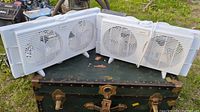 Photo shows two white window fans with dual blades placed outdoors on a metal box, one bundled with cord.