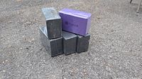 Five yoga blocks, four black and one purple with Gaiam branding, stacked outdoors on gravel surface.