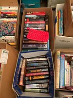 Two boxes of books containing various novels stacked with a rose-patterned zippered bag on top of one box.