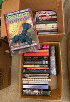 Box containing several paperback and hardcover books stacked horizontally and vertically, and a colorful tartan plush crab on top along with a Forest City Comicon event program.