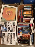 Two boxes containing paperback novels and art prints. One box contains a mix of books by Danielle Steel, Nora Roberts, and others. Includes a colorful art plaque print and a black and white small poster.