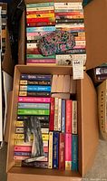 Two boxes densely packed with paperback romance novels from multiple authors with colorful spines. Makeup bag and marble horse head bookend visible in the box.