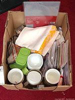 Top view of box with three white bowls, green divided plastic container, towel set with yellow border, assorted note cards, embroidered mat and other small kitchen items