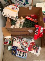 Photo showing box filled with linens, garland, Santa and angel figurines, and assorted ornaments including a tablecloth and stockings.