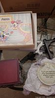 Photo showing Disney-themed photo album with Mickey and Minnie Mouse on cover, baby's musical photo box corner, baptism plaque with visible floral design and inscription, and partial view of a plate holder.