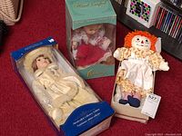 Lot of three dolls displayed together: porcelain doll in winter coat in box, musical animated doll in green box, and Raggedy Ann doll outside box.