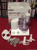 Overall view of the Braun CombiMax 600 food processor with box and various attachments laid out on red carpet.