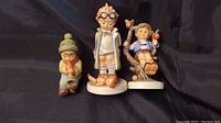 Three Hummel figurines displayed side by side: Sound the Trumpet, Doctor, and Apple Tree Boy