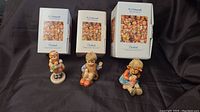 Three Hummel figurines with original boxes shown on dark fabric background