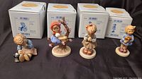 Four Hummel figurines in a row with their boxes behind them.