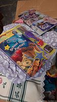 Box showing assorted linens including patterned fabric and lace-edged pieces, the Company’s Coming School Days Parties cookbook, a floral gift bag, and party accessory items such as balloons and decorations.