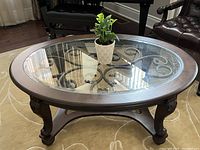 Front perspective view of the oval coffee table showing wood frame, carved legs and scrolled wrought iron base under glass top