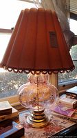 Front side view of the bubble glass pumpkin-shaped lamp showing clear glass base with bubble texture and decorative gold swirls, topped with an orange pleated fabric lampshade with dark brown fringe trim.