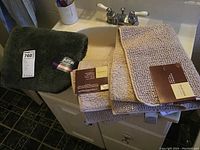 Three bathroom mats stacked on a bathroom counter: dark green plush solid mat and two beige textured mats, all with tags.