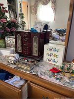 Photo showing multiple jewelry and trinket boxes on a dresser, including a dark wooden cabinet with etched glass doors, a white floral drawer box, and assorted decorative items.