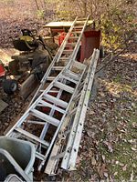Overview photo of the three aluminum ladders stacked outdoors showing extension and A-frame types amidst other yard equipment and leaves.
