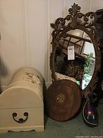 Photo of ornate framed oval wall mirror, floral painted keepsake chest, round carved wood plaque, and iridescent bottle in metal holder, showing the lot contents together.